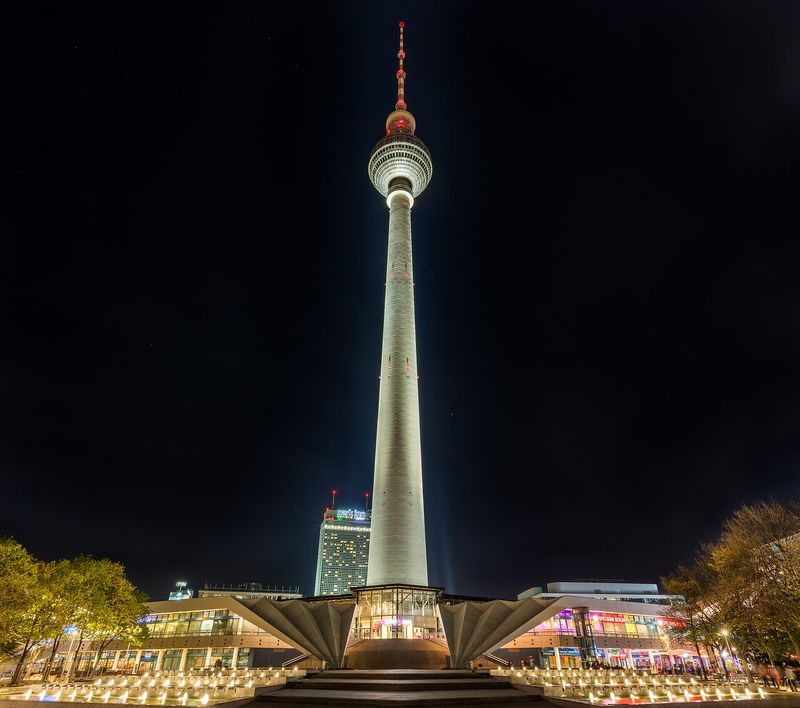 Der hell angestrahlte Fernsehturm bei Nacht, vom Fuß des Turms aus gesehen, mit dem gefalteten Dach des Sockelbaus im Vordergrund
