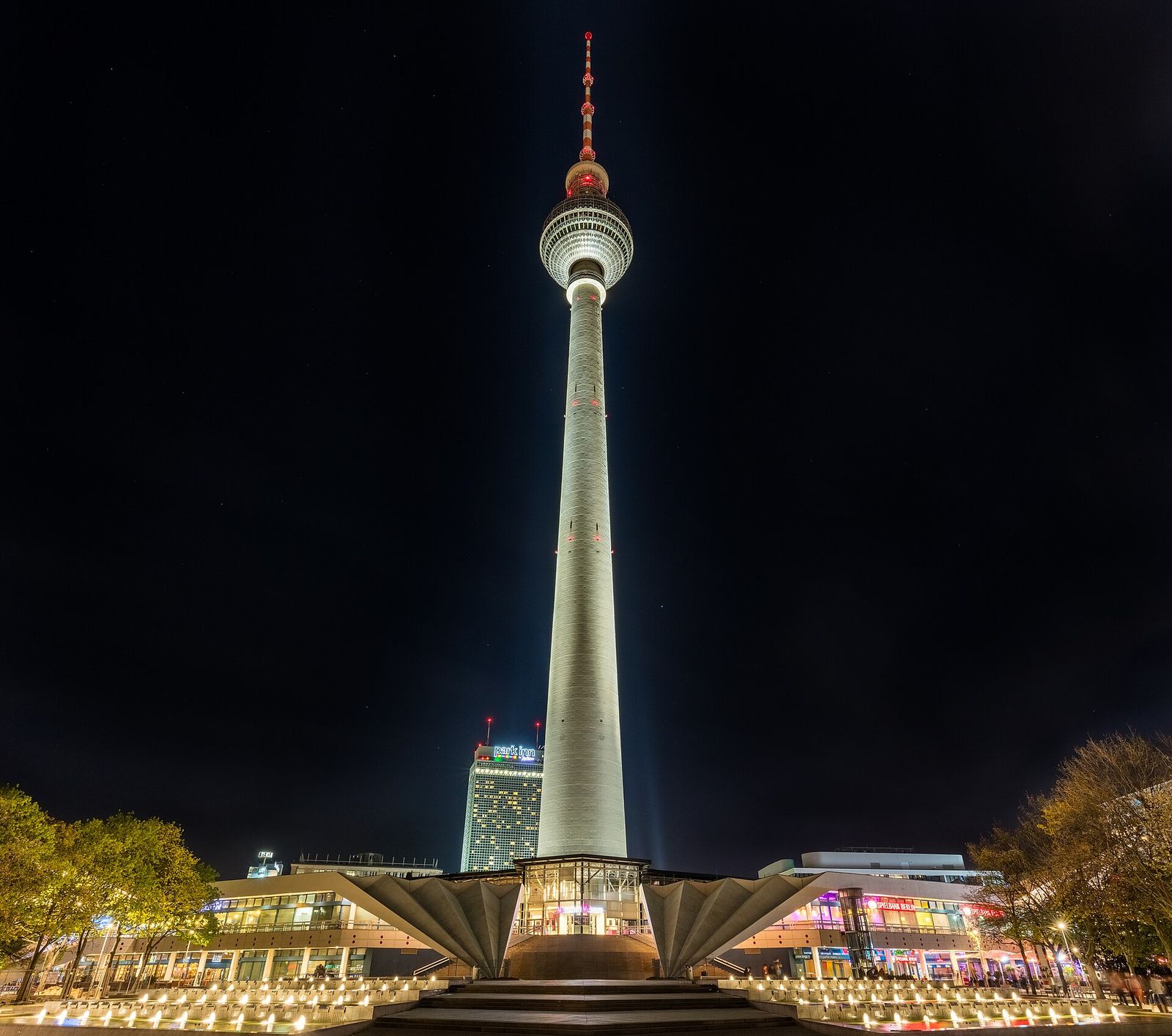 Der hell angestrahlte Fernsehturm bei Nacht, vom Fuß des Turms aus gesehen, mit dem gefalteten Dach des Sockelbaus im Vordergrund