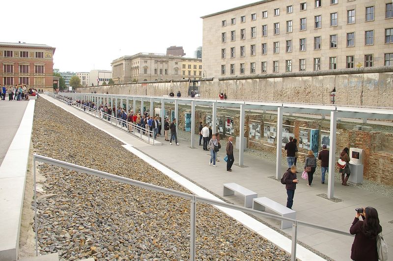 Freigelegte Kellermauern mit Ausstellungstafeln im Graben der Topographie des Terrors, darüber ein erhaltenes Stück der Berliner Mauer an der Niederkirchnerstraße