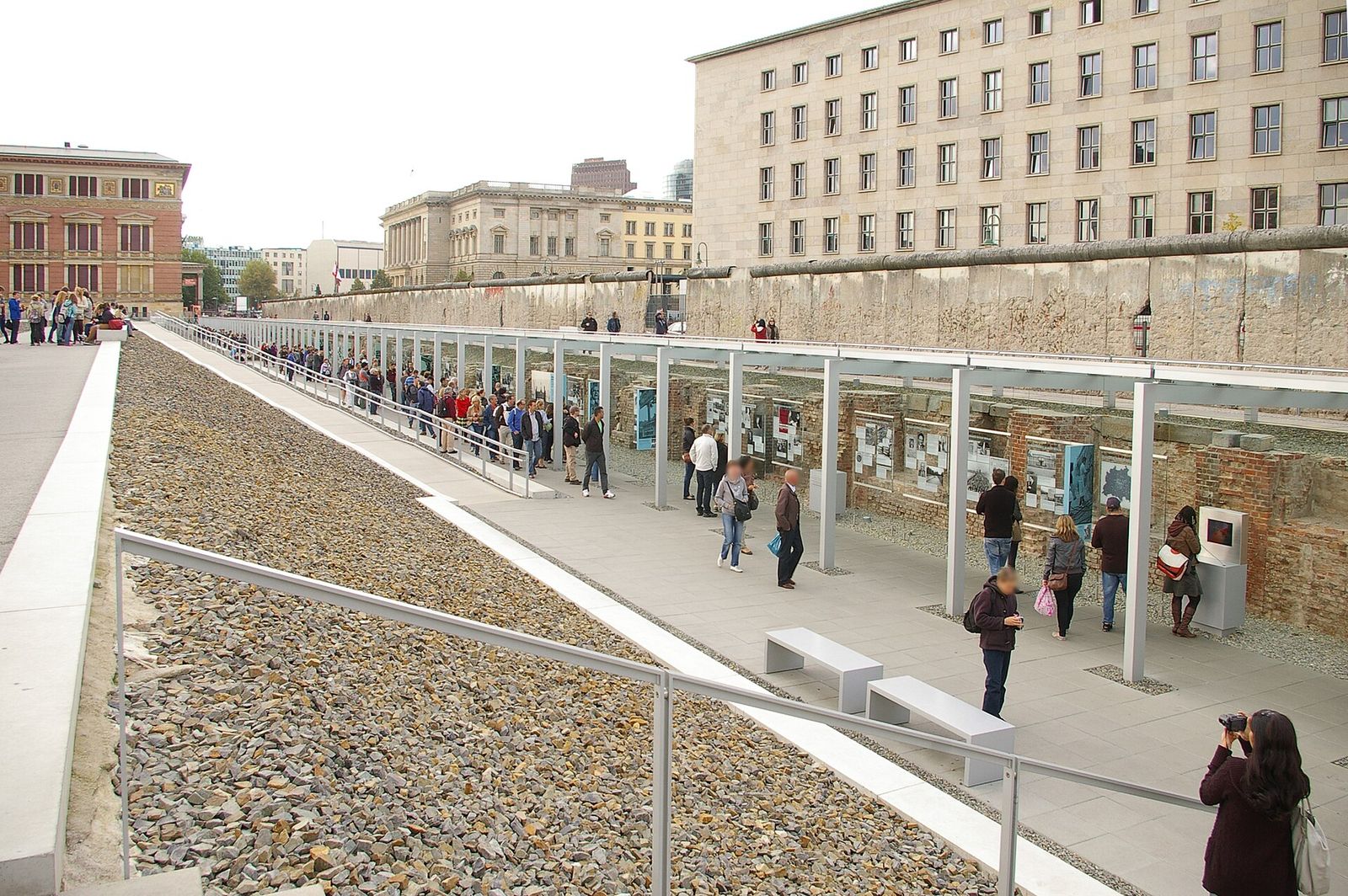Freigelegte Kellermauern mit Ausstellungstafeln im Graben der Topographie des Terrors, darüber ein erhaltenes Stück der Berliner Mauer an der Niederkirchnerstraße