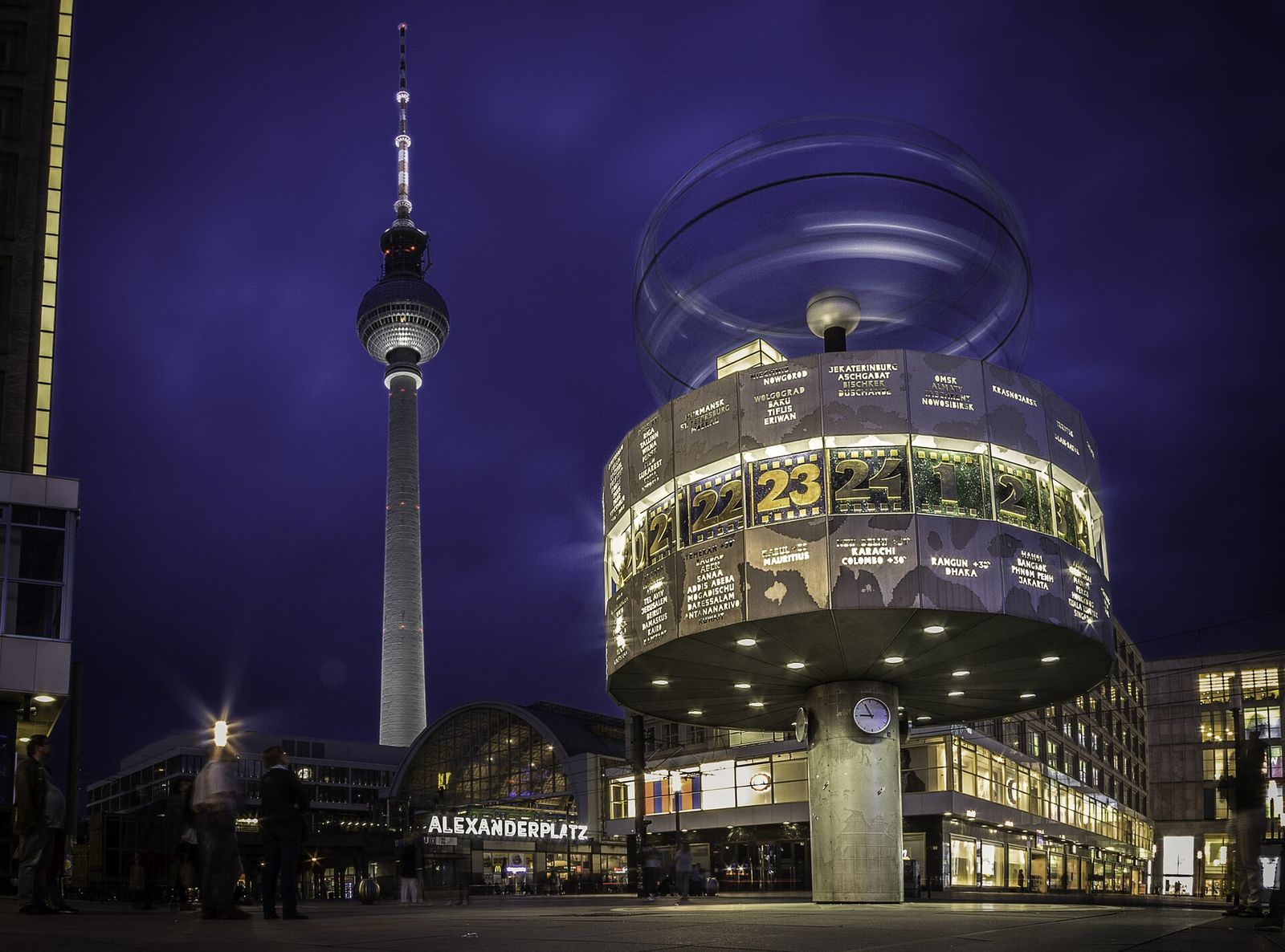 Die beleuchtete Weltzeituhr auf dem Alexanderplatz am Abend, dahinter der Fernsehturm und der Bahnhof Alexanderplatz